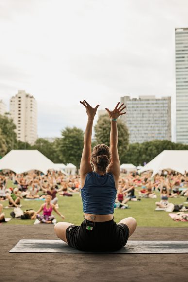 Frau im Vordergrund, die Yoga macht. Mehrere Personen im Hintergrund auf der Wiese, die die Übung nachmachen.